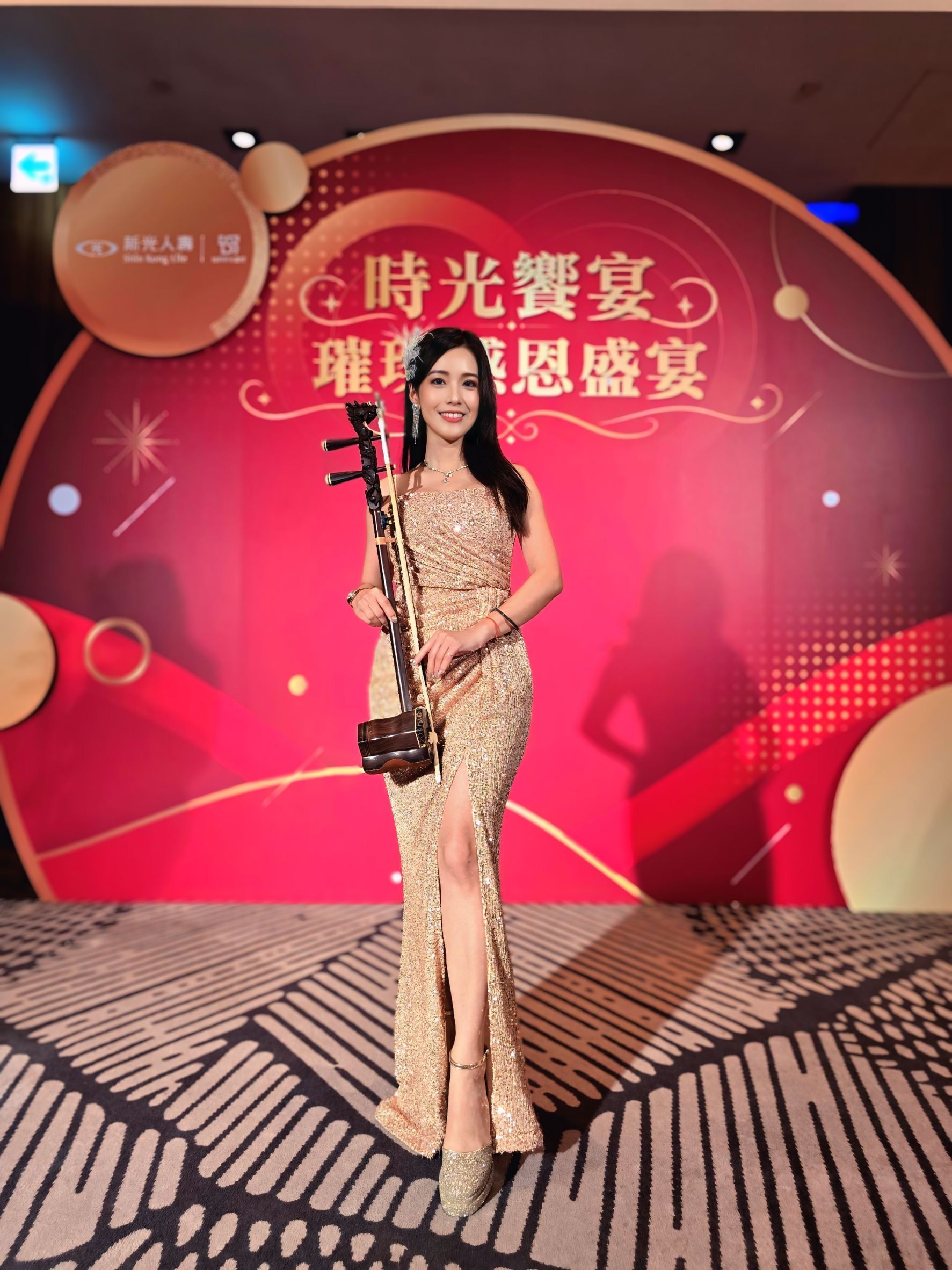 胡琴姐姐身穿金色禮服，於新光人壽感恩盛宴背板前持二胡
